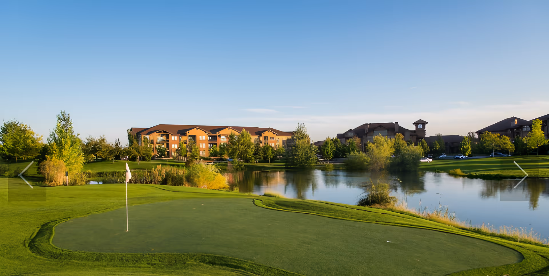 A golf green with a flag beside a pond, trees, and a senior living complex in the background under a clear blue sky.