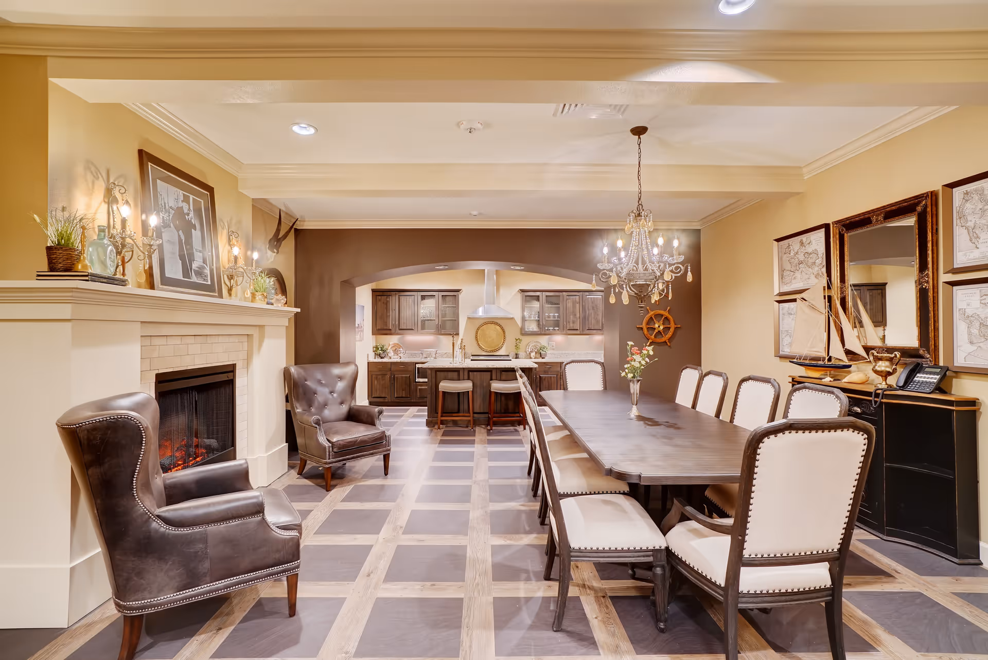 Elegant dining room with a long wooden table and upholstered chairs, a chandelier overhead, leather armchairs by a fireplace, and a kitchen visible in the background.