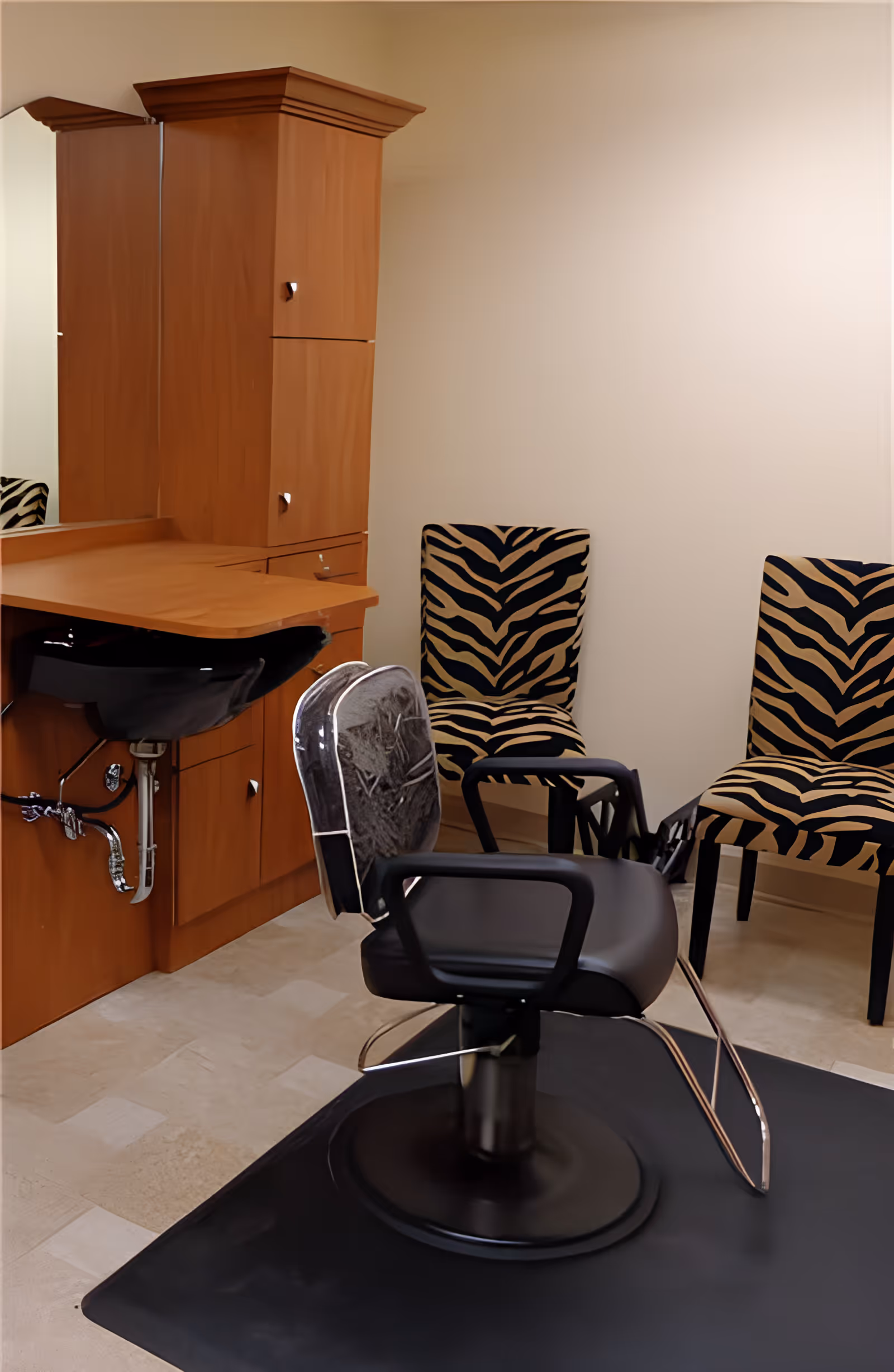 Interior view of a salon area in a senior living community featuring a black salon chair with a protective cover, a wooden vanity with a black sink, and two zebra-patterned chairs against a plain wall.