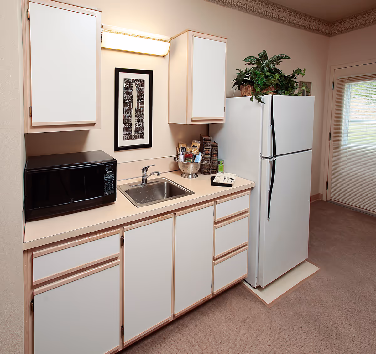 A small kitchen area with white cabinets, a stainless steel sink, a black microwave on the counter, and a white refrigerator with a potted plant on top. There is a framed word art hanging on the wall above the sink and a door with blinds to the right.