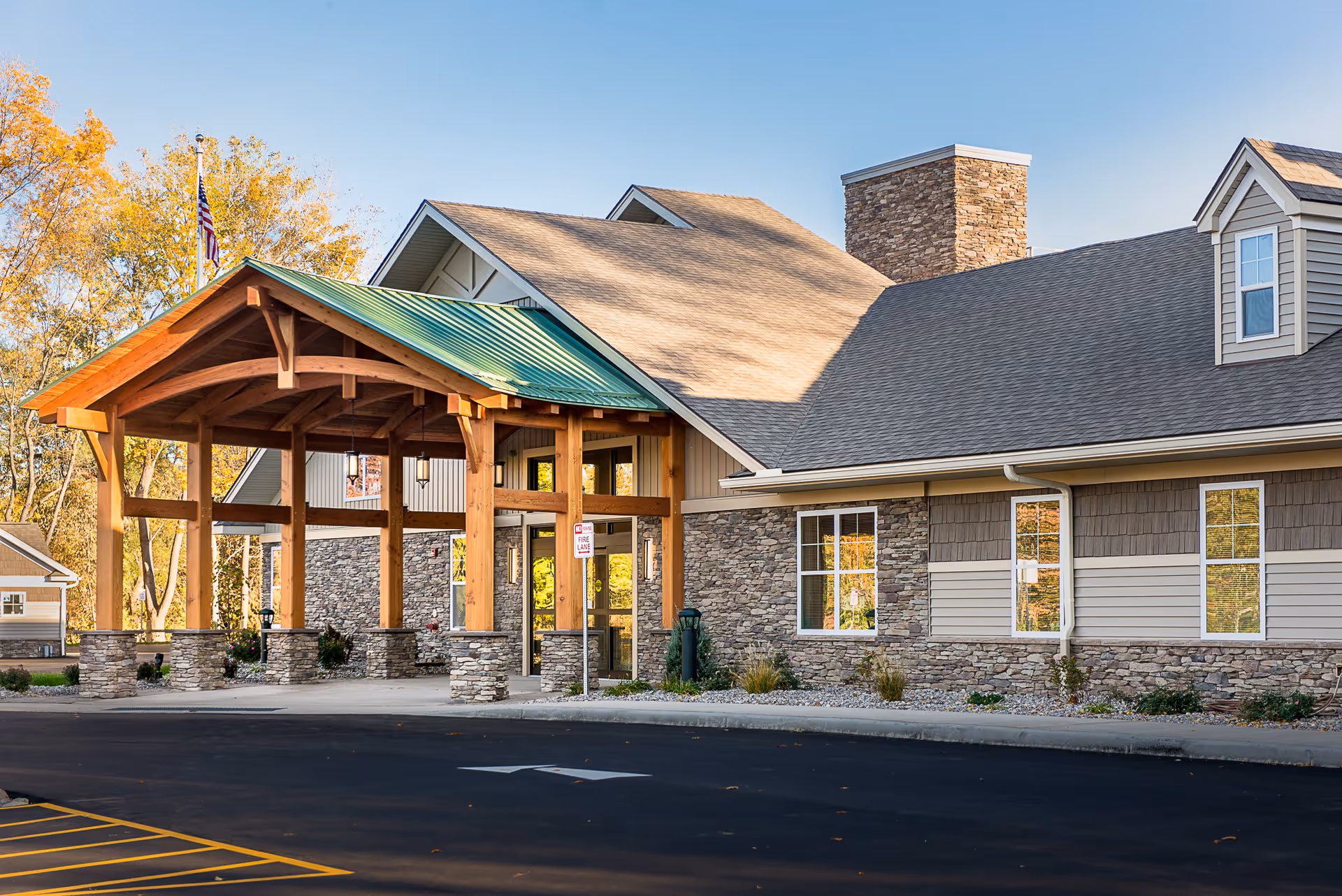 Exterior view of a senior living facility building with a covered entrance supported by wooden beams and stone pillars. The building has a stone and siding facade with multiple windows and a chimney. Trees with autumn foliage are visible in the background under a clear blue sky.