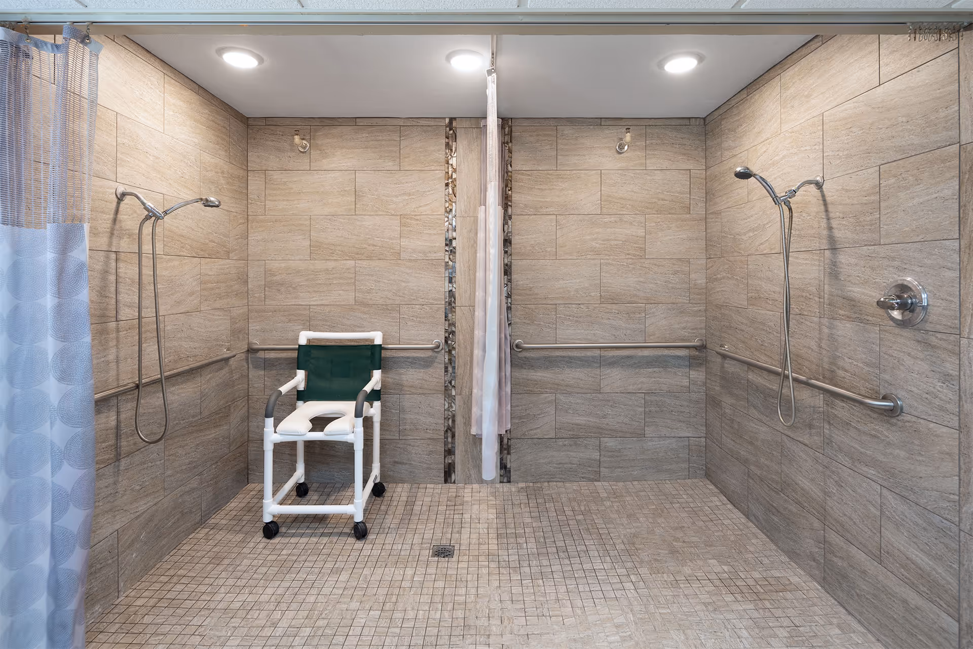 A spacious accessible shower area with two shower heads on opposite walls, grab bars along the tiled walls, and a white shower chair with green fabric seating positioned on the left side. The floor and walls are covered with beige tiles, and a shower curtain is partially visible on the left side.