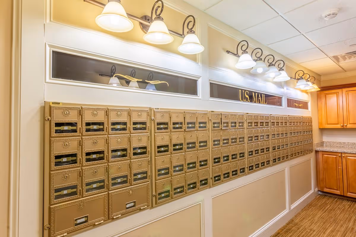 Interior view of a mailroom with a wall of gold-colored mailboxes labeled U.S. MAIL, illuminated by multiple wall-mounted light fixtures above. To the right, there are wooden cabinets and a granite countertop.