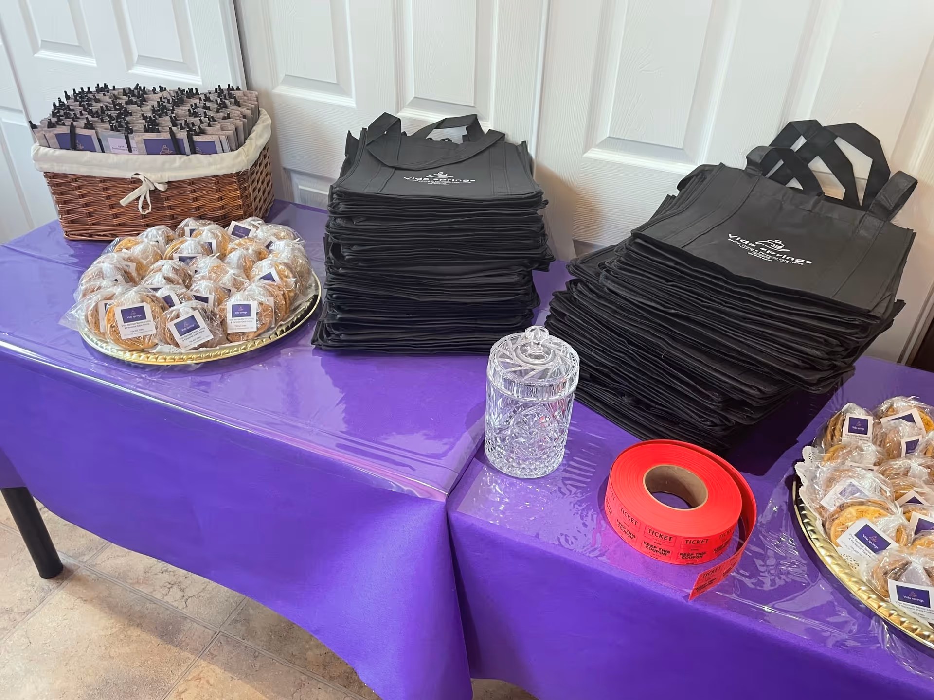 Table with purple tablecloth holding stacks of black tote bags, trays of wrapped cookies, a glass jar, and a roll of red raffle tickets.