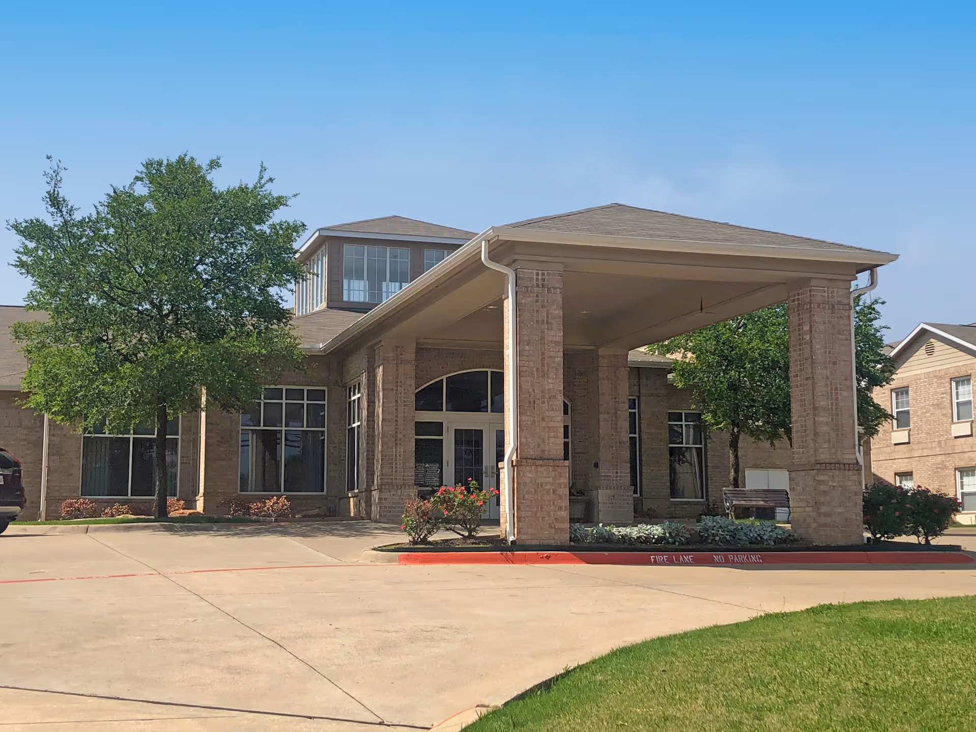 Exterior view of a brick building with a covered entrance supported by four large pillars. There are trees and shrubs around the building, a clear blue sky, and a driveway with a red curb marked 'FIRE LANE NO PARKING'.