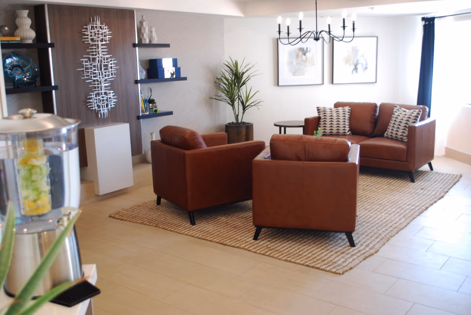 A modern living room with two brown leather armchairs and a matching sofa arranged on a woven rug. The sofa has two patterned pillows. Behind the sofa are two framed abstract artworks on the wall and a black chandelier hanging from the ceiling. To the left, there are decorative shelves with vases and a metallic wall sculpture. A water dispenser with fruit-infused water is partially visible in the foreground.