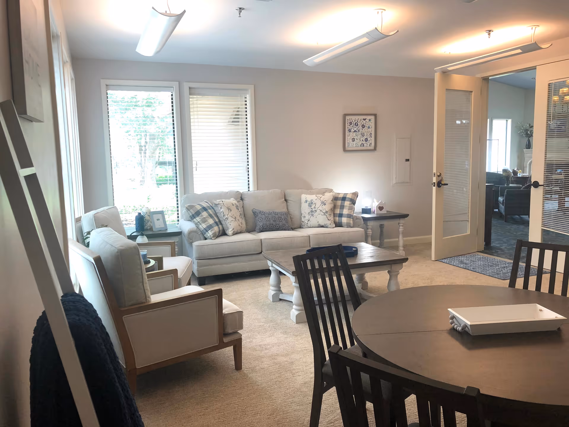 A cozy living room area with a beige sofa adorned with patterned pillows, two beige armchairs, a wooden coffee table, and a round dining table with chairs. The room has large windows with blinds, soft lighting from ceiling fixtures, and an open door leading to another room.