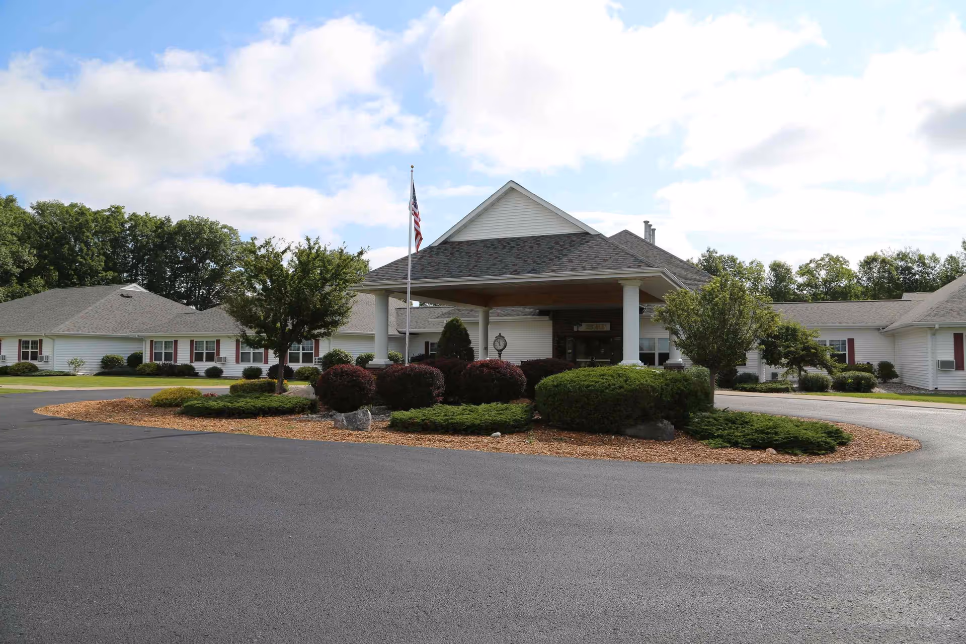 Single-story white senior living building with a covered porte-cochère entrance, circular driveway, landscaped shrubs, and an American flag.