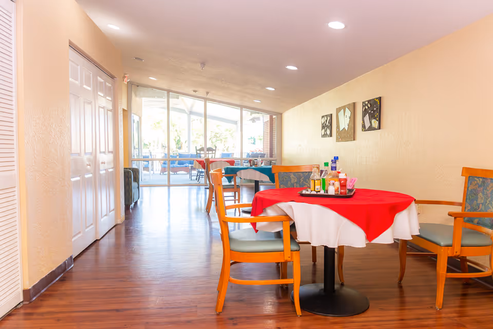 Interior view of a dining area in Bethesda on Turkey Creek with wooden chairs around tables covered with white and red tablecloths. Condiments are placed on a tray on the nearest table. The room has wooden flooring, beige walls with decorative artwork, and large windows at the far end letting in natural light.