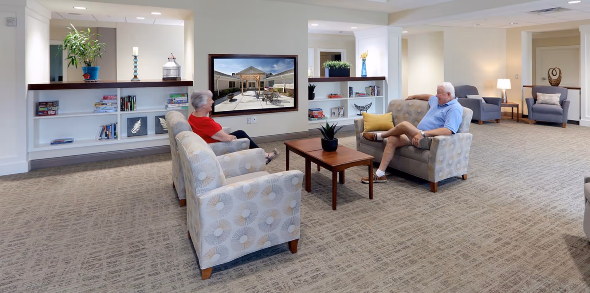 A spacious and well-lit senior living facility lounge area with two elderly people sitting on patterned armchairs around a wooden coffee table with a small plant. The room features built-in white shelves with books and decorative items, a large wall-mounted TV displaying an outdoor patio scene, and additional seating with armchairs and side tables with lamps in the background.