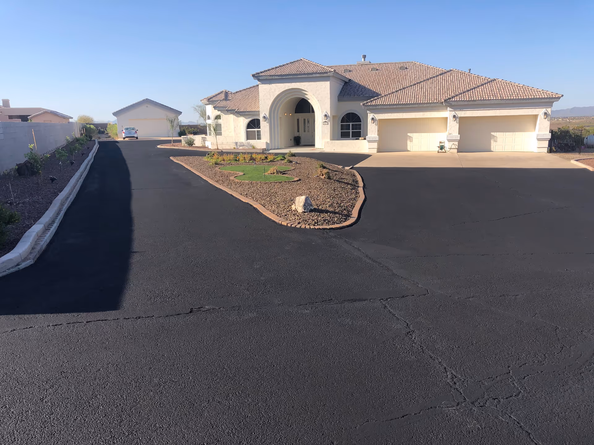 A large single-story house with a tiled roof and an arched entrance, surrounded by a freshly paved black asphalt driveway. There is a landscaped area with rocks and small plants in front of the house, and a detached garage is visible in the background. The sky is clear and blue.