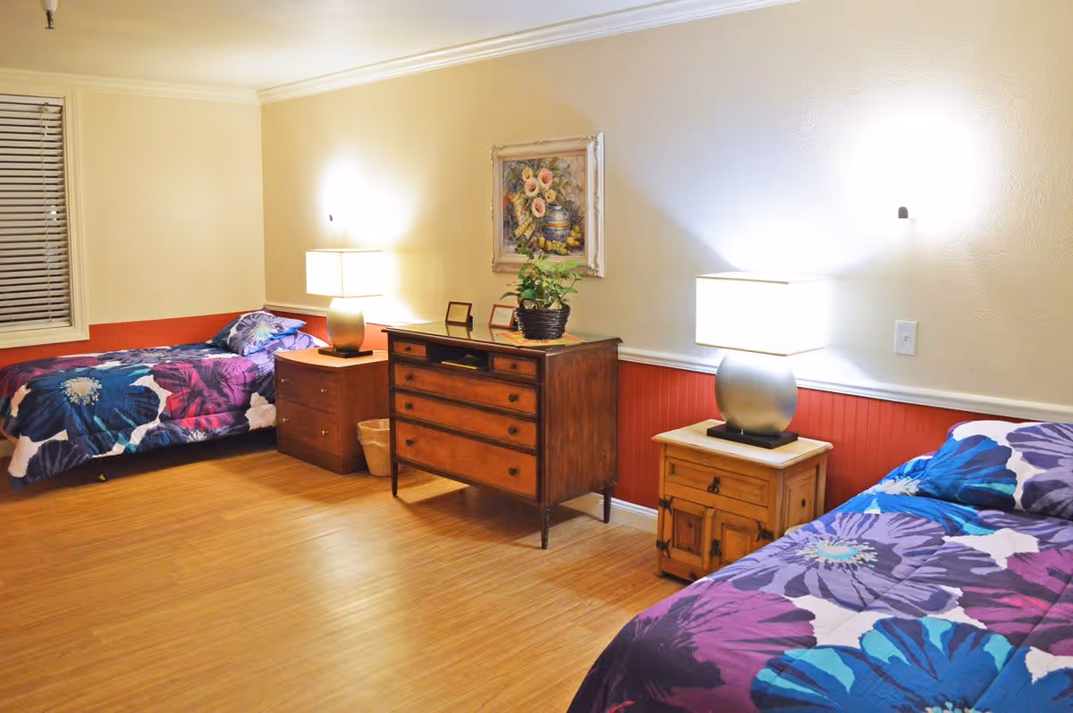 A cozy bedroom with two single beds, each covered with colorful floral bedspreads in shades of purple, blue, and white. Between the beds are two wooden nightstands, each with a modern table lamp. A wooden dresser with drawers is placed against the wall, decorated with a potted plant and framed pictures. Above the dresser hangs a framed painting of flowers and fruit. The room has light-colored walls with a red wainscoting and wooden flooring.