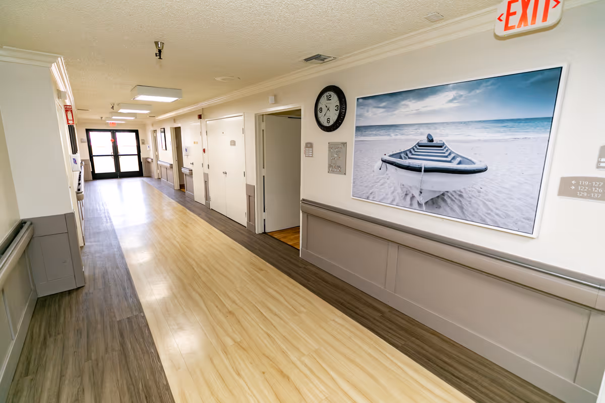 A clean, well-lit hallway in a healthcare facility with light wood and gray flooring, white walls, a large framed photo of a boat on a beach, a wall clock, and an exit sign above a door at the end of the corridor.