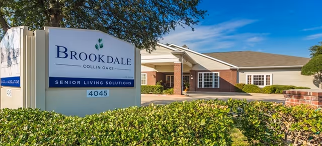 Exterior view of Brookdale Collin Oaks senior living facility with a large sign in front displaying the name and address, surrounded by greenery and a clear blue sky.