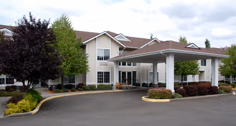 Two-story senior living building with a covered entrance/porte-cochère, landscaped shrubs and trees, and a paved driveway.
