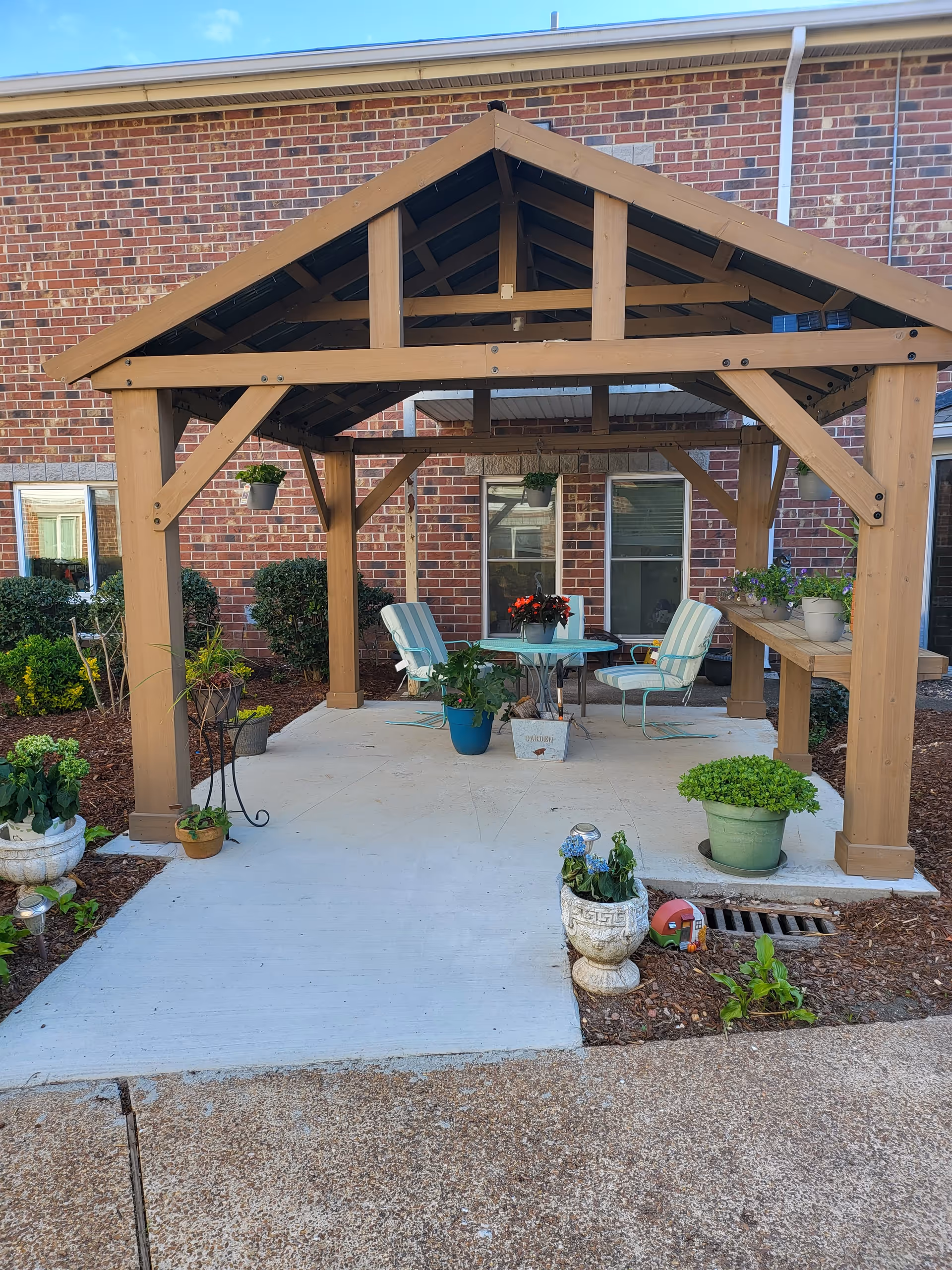 Outdoor covered patio area with a wooden pergola structure, featuring a round glass table with four striped cushioned chairs. Various potted plants and flowers are placed around the patio and hanging from the pergola. The background shows a brick building with windows and some shrubbery.