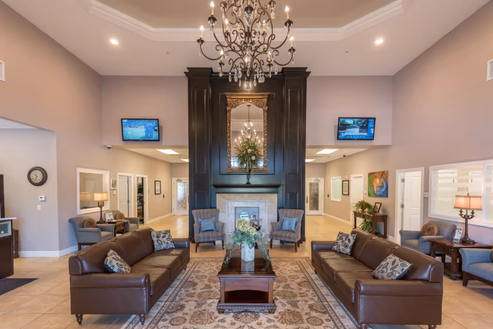 A spacious and elegant living room area with two brown leather sofas facing each other, a glass coffee table with a floral arrangement in the center, and two armchairs near a large black fireplace with a decorative mirror and flowers above it. The room features a chandelier hanging from the ceiling, two wall-mounted TVs, and soft lighting from table lamps on side tables. The walls are painted in a light beige color, and the floor is tiled with a patterned area rug in the seating area.