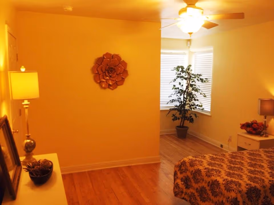 Warmly lit bedroom with hardwood floors, a bed and nightstand, lamps, wall decor, and a potted plant near a bay window.