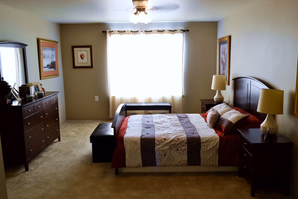 A well-lit bedroom with a large window covered by light curtains. The room features a bed with a patterned quilt and multiple pillows, two bedside tables each with a lamp, a dark wooden dresser with a mirror, and framed artwork on the walls. The carpet is beige, and there is a ceiling fan with lights above.