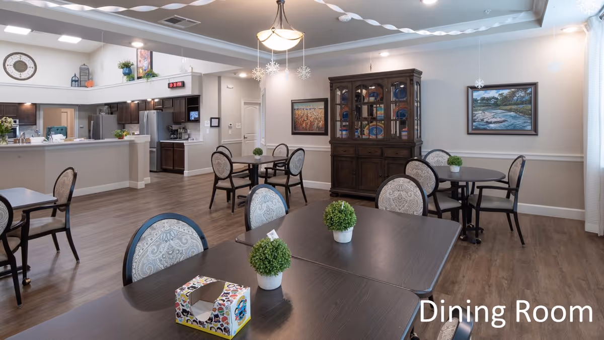 A spacious dining room with several dark wood tables and cushioned chairs arranged neatly. Small potted plants are placed on the tables. The room features wood flooring, a china cabinet filled with plates, framed paintings on the walls, and a kitchen area visible in the background. Ceiling lights and hanging snowflake decorations add to the ambiance.