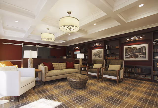 A cozy living room with a beige sofa, two armchairs, a white armchair, and a round wooden coffee table on a plaid carpet. The walls are painted dark red with built-in bookshelves filled with books and framed artwork. Two large ceiling lights and two table lamps provide warm lighting.