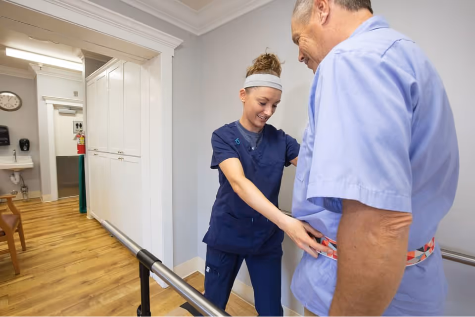 A healthcare worker in navy scrubs is assisting an elderly man in a light blue shirt with a gait belt in a hallway with wooden flooring and white walls. There is a handrail along the wall and a sink with a clock and fire extinguisher visible in the background.