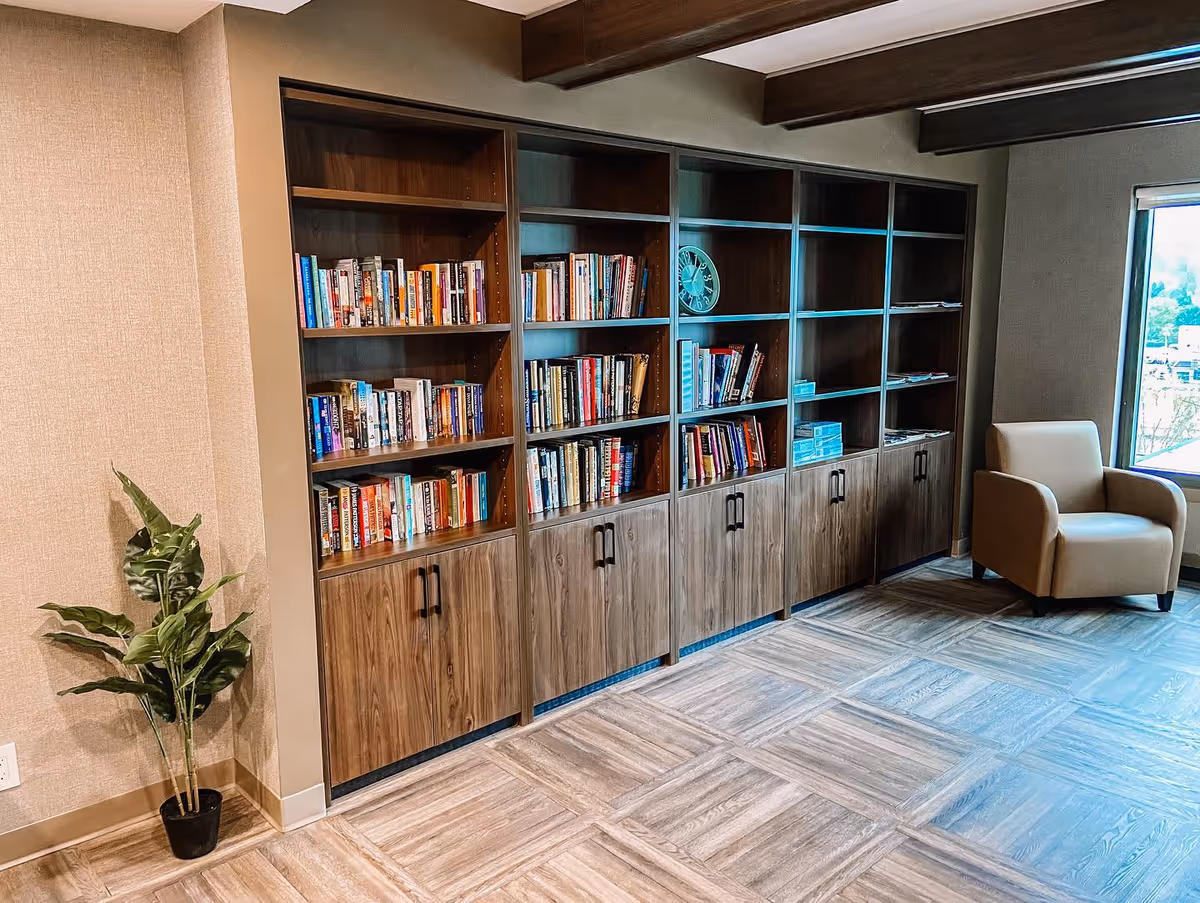 A cozy reading area in a senior living facility featuring a large wooden bookshelf filled with books, a beige armchair near a window, and a potted plant in the corner. The room has tiled flooring and wooden ceiling beams.