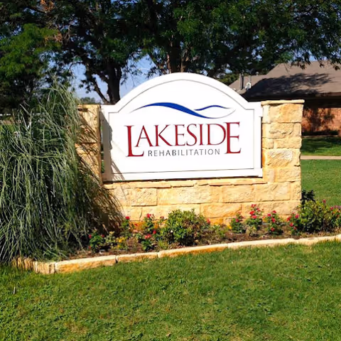 A stone sign with the words 'Lakeside Rehabilitation' in red and blue lettering, surrounded by green grass, plants, and trees in an outdoor setting.