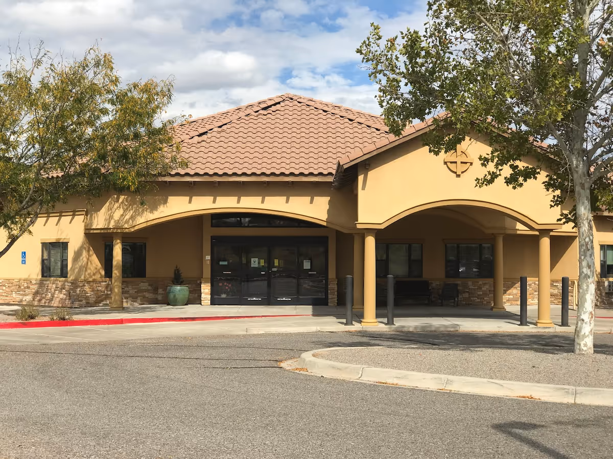 Front exterior view of a single-story building with a tiled roof and beige walls, featuring a covered entrance supported by columns. There are trees on either side of the entrance and a parking area in front.