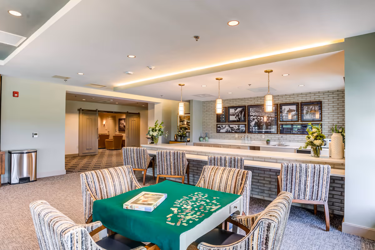 A well-lit common area with a table covered in a green cloth and scattered puzzle pieces, surrounded by six striped upholstered chairs. In the background, there is a long counter with four matching chairs, pendant lights hanging from the ceiling, and framed black and white photos on a brick wall. The room has a carpeted floor and an open doorway leading to another seating area.