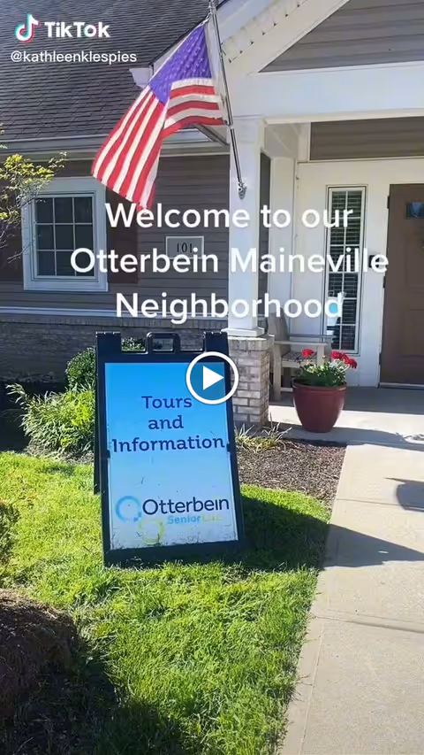 Front entrance of Otterbein Maineville SeniorLife with an American flag, potted flowers, and a sidewalk sign reading "Tours and Information".
