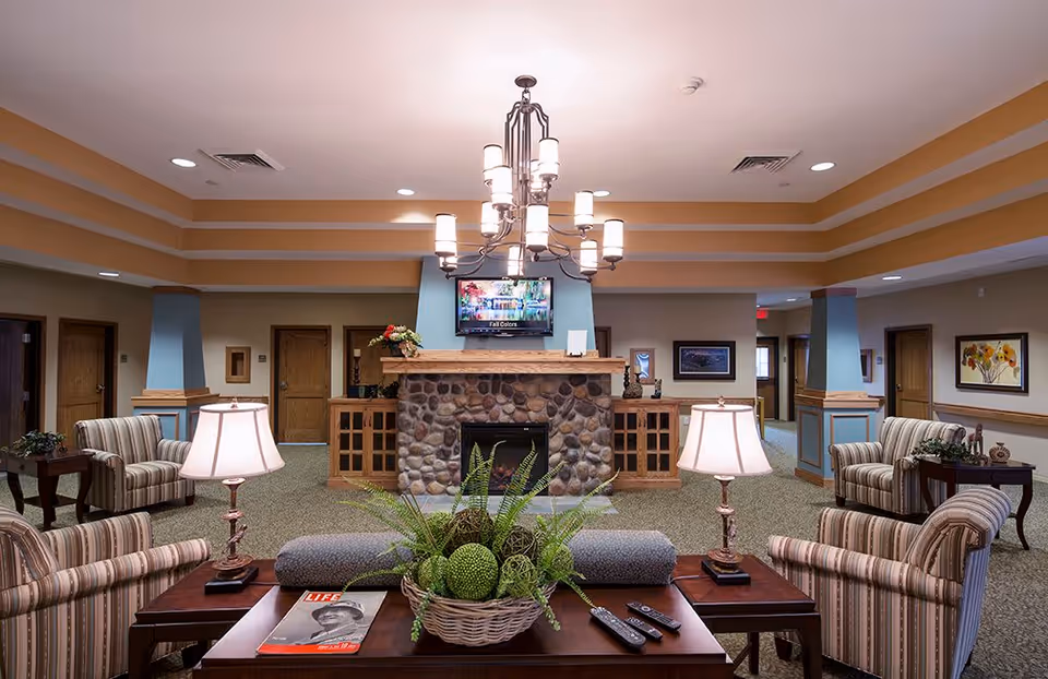 A cozy communal living room with striped armchairs, a central stone fireplace topped by a TV and chandelier, and a coffee table with a plant and magazines.
