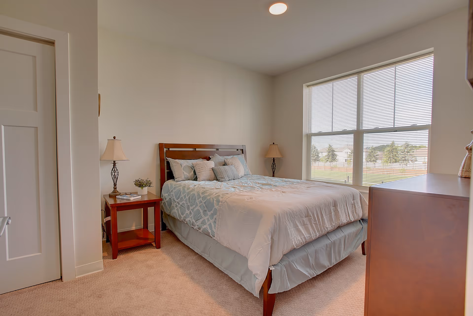 A bright bedroom with a large window covered by blinds, a bed with blue and white bedding and multiple pillows, two bedside tables each with a lamp, a small plant, and a dresser on the right side.