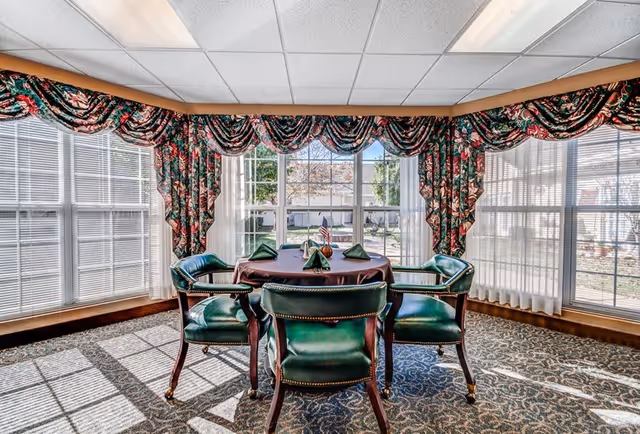 A bright dining area with large windows covered by floral valances and sheer curtains. In the center, a round table with a dark tablecloth is set with folded green napkins and surrounded by four green leather chairs with wooden legs. Sunlight streams through the windows, casting shadows on the patterned carpeted floor.