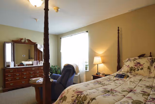 A cozy bedroom with a four-poster bed covered in floral bedding, a wooden dresser with a mirror, a blue recliner chair, a small wooden side table, a lamp on a nightstand, and a window with sheer curtains letting in natural light.