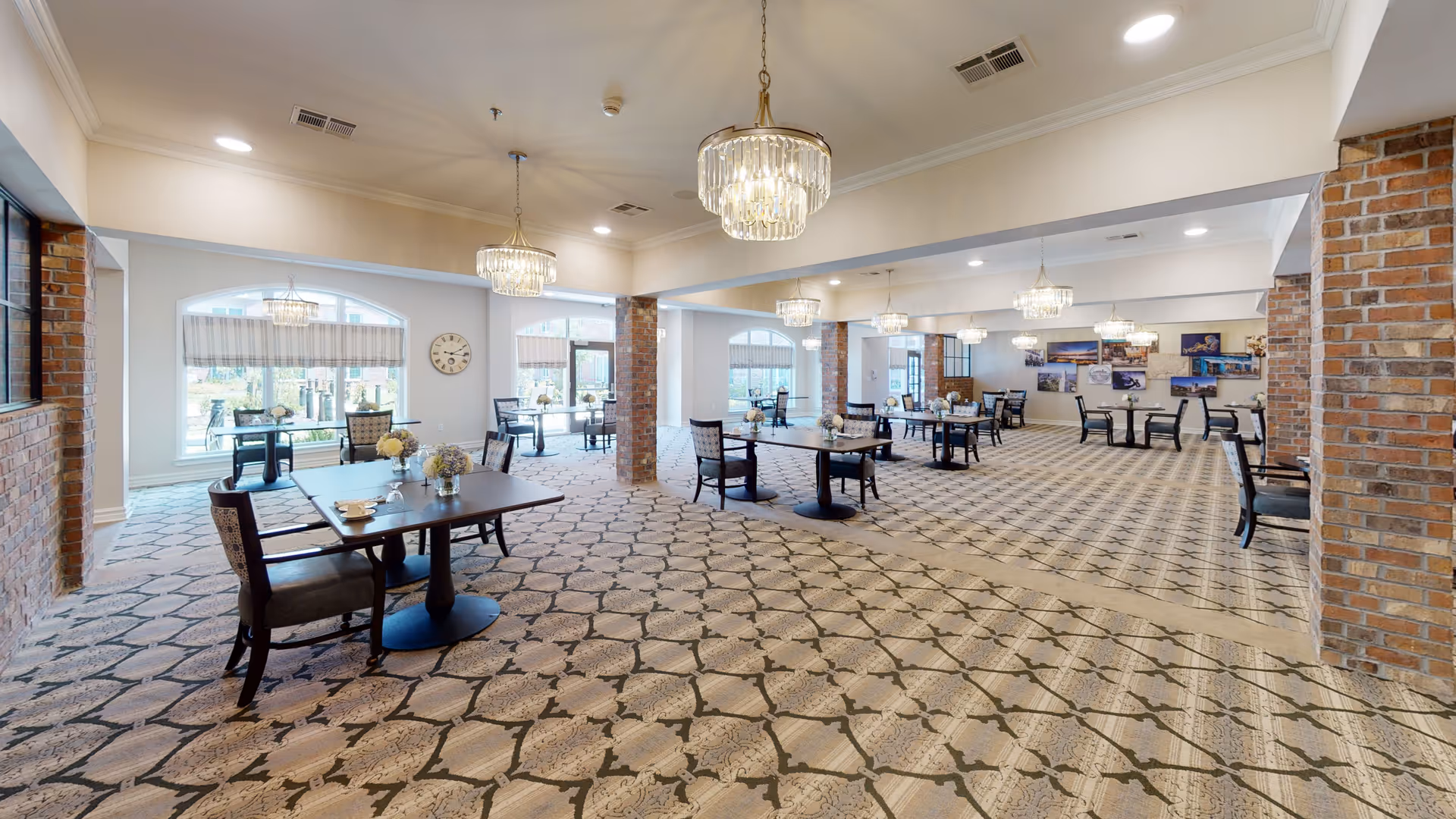 Large dining room with multiple tables and chairs, crystal chandeliers, patterned carpet, and exposed brick columns.