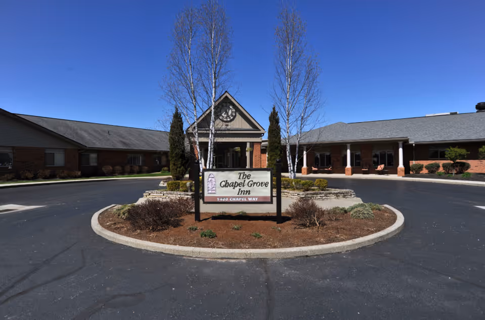 Entrance to The Chapel Grove Inn showing the building's front, central sign, circular driveway, and landscaping under a clear blue sky.