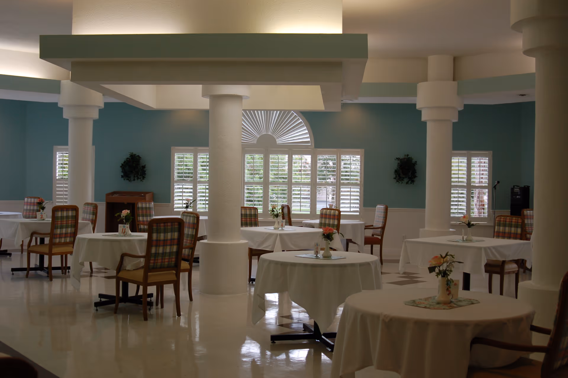 A dining room with round tables covered in white tablecloths, each decorated with a small vase of flowers. The room has teal walls, white pillars, and windows with white shutters letting in natural light. Chairs with plaid upholstery are arranged around the tables.