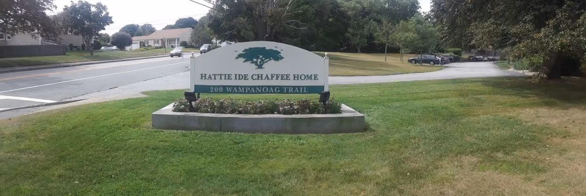 A large sign on a grassy area near a road that reads 'Hattie Ide Chaffee Home, 200 Wampanoag Trail' with trees, parked cars, and houses in the background.