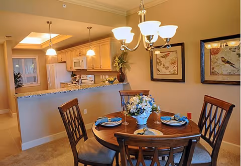 A cozy dining area with a round wooden table set for four, featuring blue napkins and a floral centerpiece. The dining area is adjacent to a kitchen with light wood cabinets, a granite countertop, and white appliances including a refrigerator, microwave, and stove. Two pendant lights hang over the kitchen counter, and a chandelier with five lights hangs above the dining table. The walls are decorated with framed bird artwork.