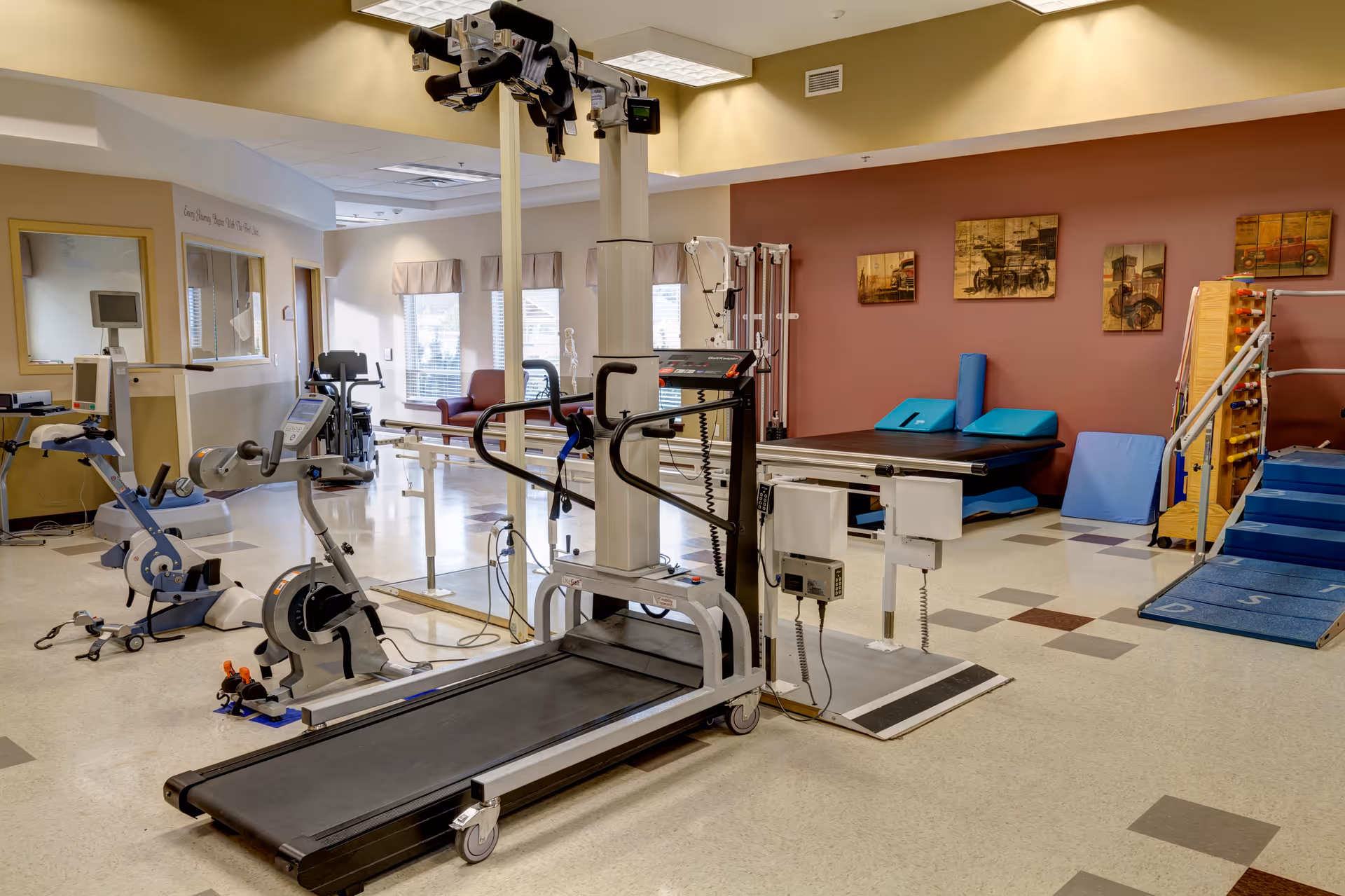 A rehabilitation or physical therapy room with various exercise and therapy equipment including a treadmill, stationary bikes, parallel bars, therapy mats, and a ramp. The room has a warm color scheme with beige and brown walls, and framed pictures on the wall.