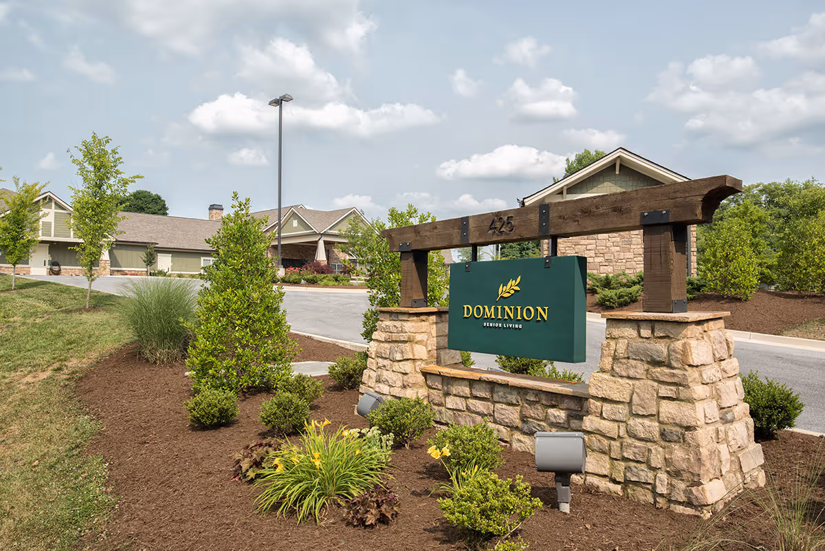 Outdoor view of Dominion Senior Living of Bristol showing a stone and wood sign with the facility name and address number 425, surrounded by landscaped greenery and bushes, with the building and a driveway in the background under a partly cloudy sky.