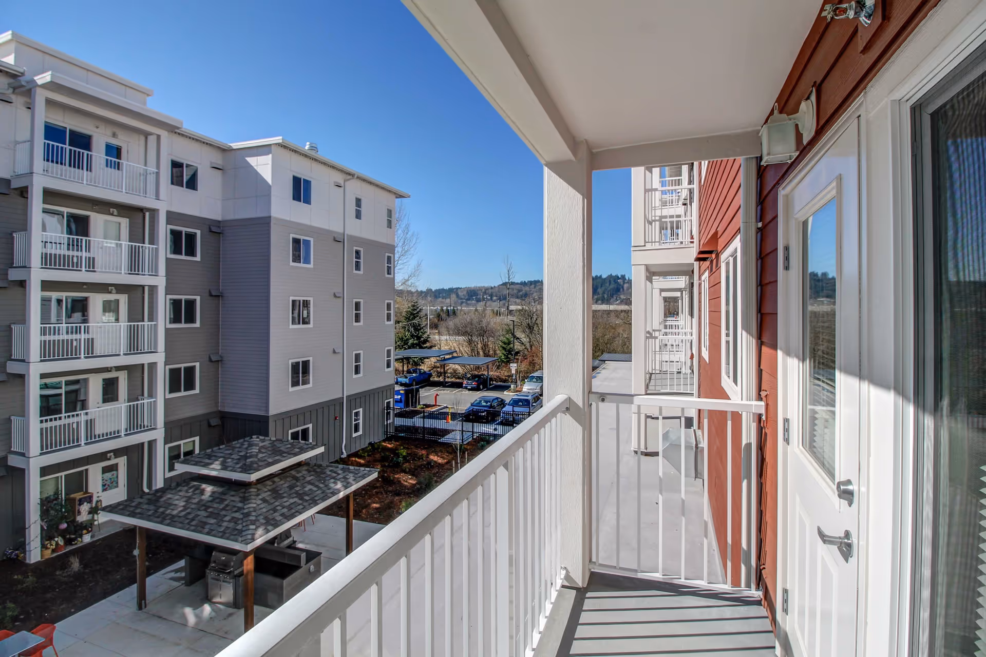 View from a covered apartment balcony overlooking neighboring multi-story residential buildings, a parking area and distant trees under a clear blue sky.