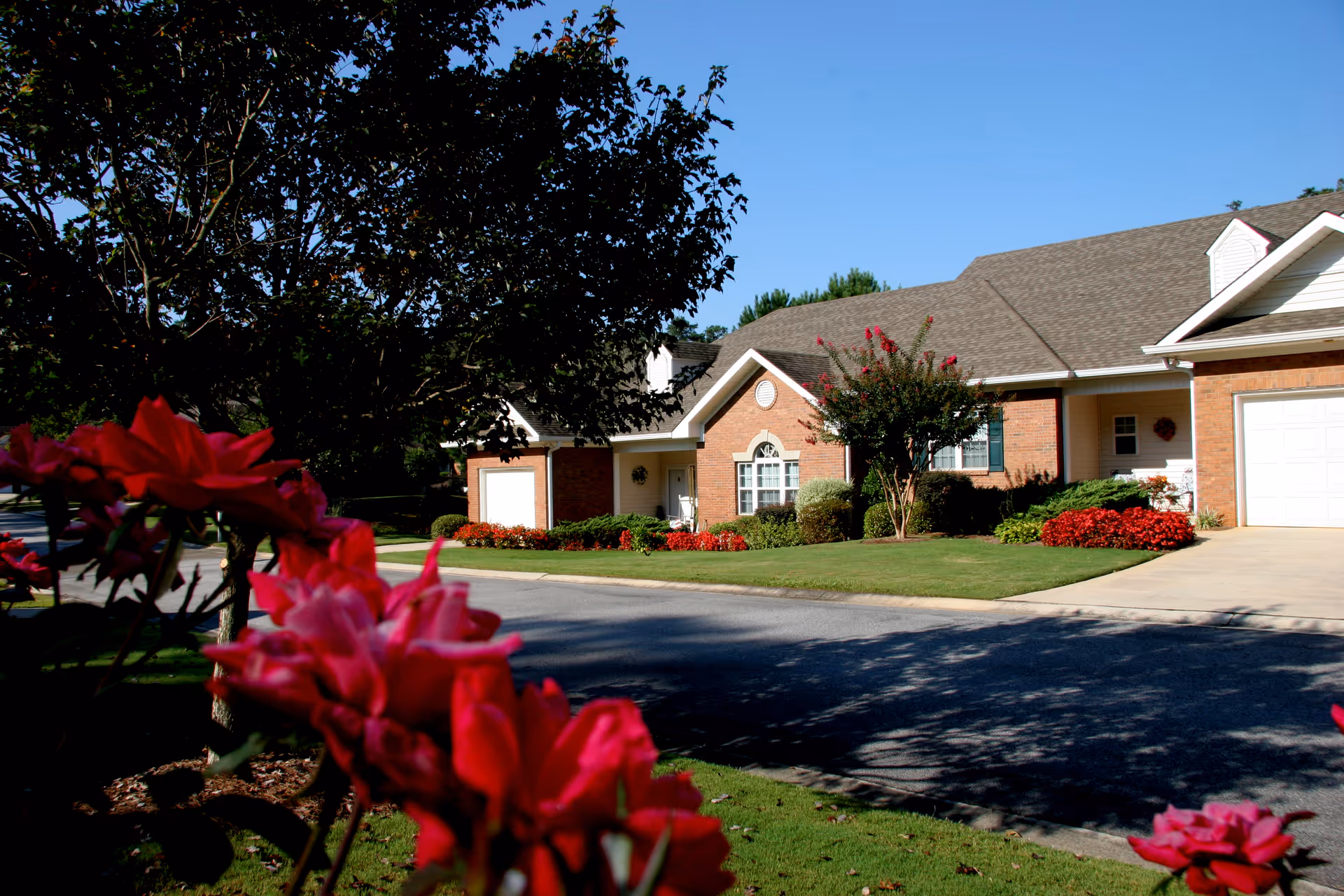 Front exterior of single-story brick cottages with garages, landscaped lawn and red flowers in the foreground under a clear blue sky.