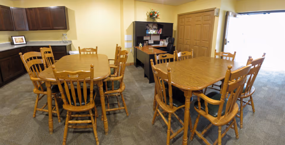 Interior room with two wooden tables surrounded by wooden chairs with green cushions. There is a countertop with cabinets on the left side and a desk with a chair and shelving unit in the back corner. The room has beige walls and carpeted floor, with a large window or door letting in bright light on the right side.