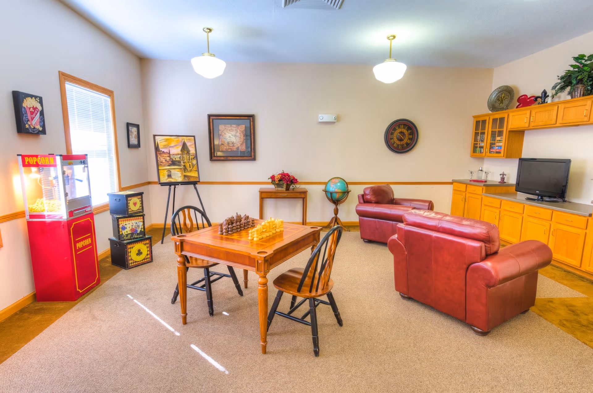 A cozy common area with a wooden chess table and two chairs in the center, two red leather armchairs, a popcorn machine near a window, framed artwork on the walls, a globe on a stand, and a TV on a wooden cabinet with shelves and plants on top.