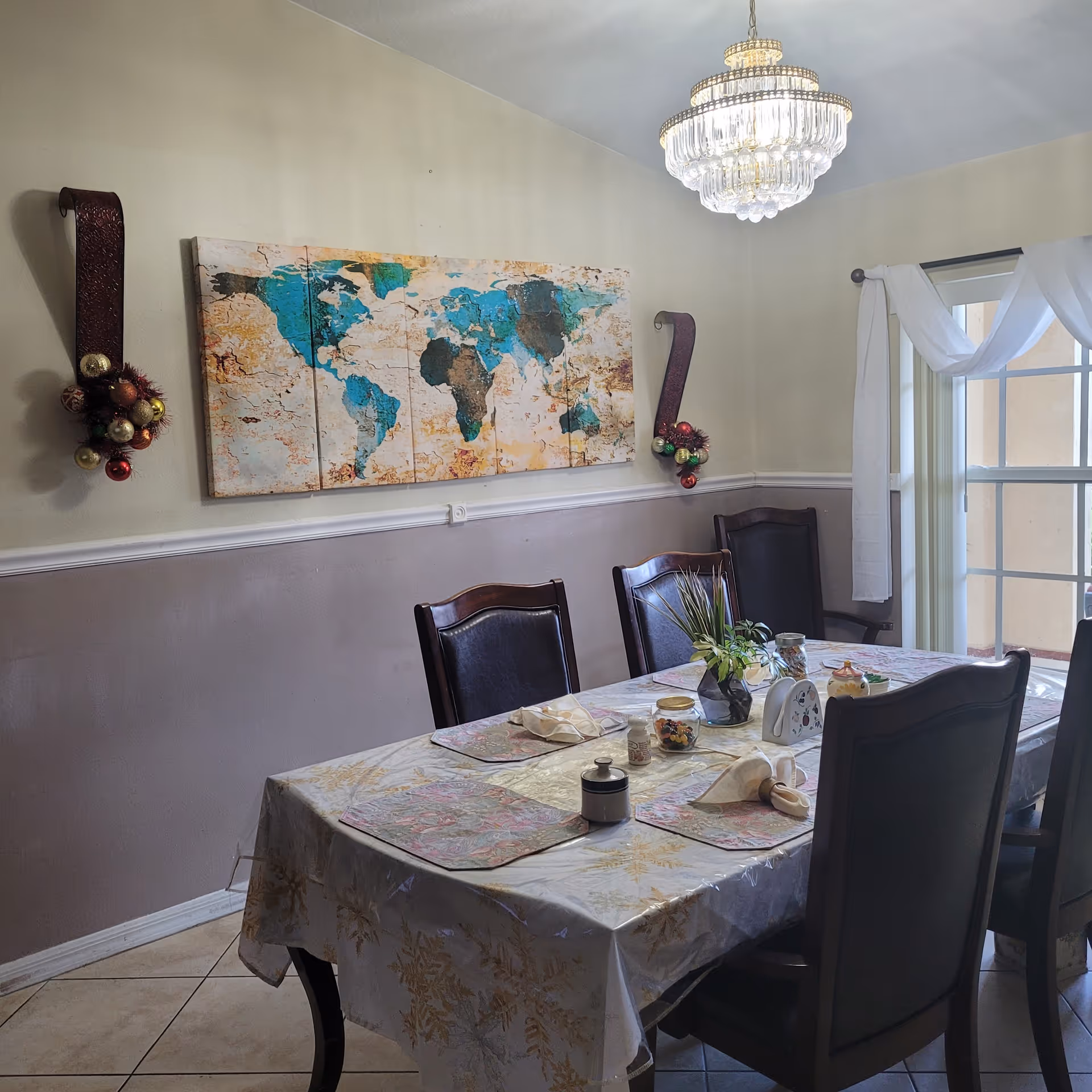 A dining room with a rectangular table covered with a white and gold patterned tablecloth and four dark wooden chairs with black cushions. The table is set with placemats, napkins, a small plant centerpiece, and various small containers. On the wall above the table is a large world map artwork divided into panels. Two decorative wall sconces with Christmas ornaments flank the map. A chandelier hangs from the ceiling, and a window with white curtains is visible on the right side.