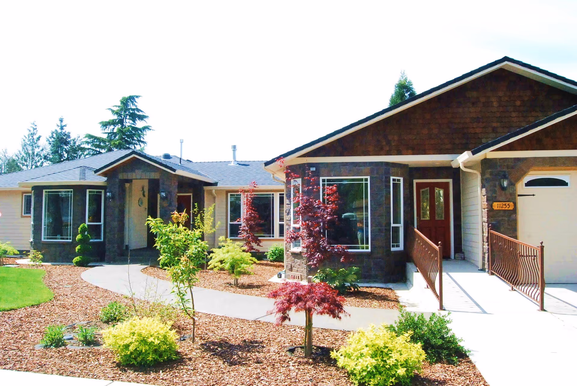 Front exterior view of a single-story senior living facility named Tigard Garden Senior Home, featuring a well-maintained garden with various shrubs and small trees, a curved concrete walkway leading to the entrance, and a ramp with railings beside the front door.