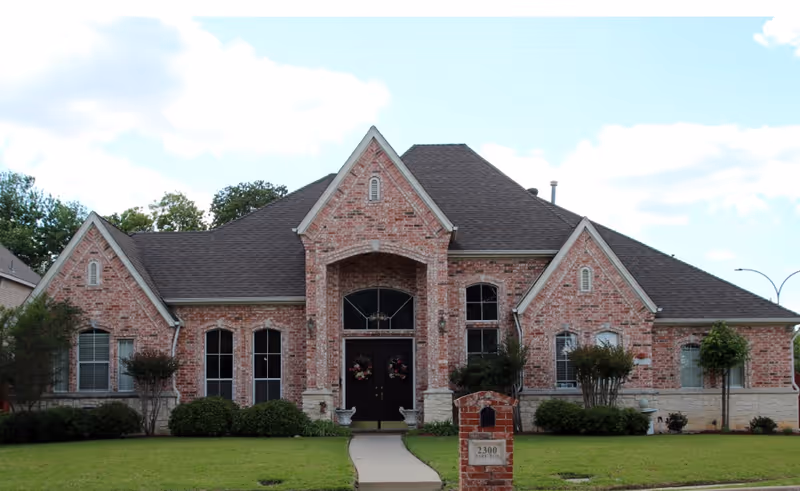 Front view of a brick, single-story house-like building with a central arched entrance, symmetrical windows, a lawn, and a mailbox.