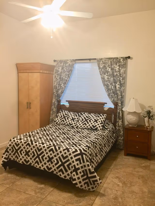 A bedroom with a double bed covered in a black and white geometric patterned bedspread and matching pillows. Behind the bed is a window with closed blinds and floral curtains. To the left of the bed is a wooden wardrobe, and to the right is a wooden nightstand with a white lamp and a small vase of flowers. The floor is tiled, and a ceiling fan with a light is visible above.